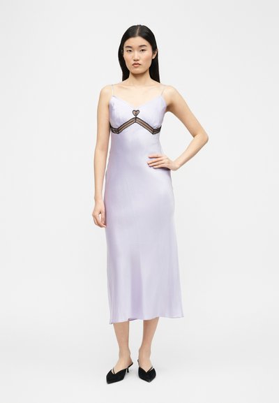 Γυναίκα με ίσια μαύρα μαλλιά φοράει λιλά σατινέ λιτό φόρεμα με λεπτομέρεια μαύρης δαντέλας σε σχήμα καρδιάς και μαύρα μυτερά mules, στέκεται μπροστά σε λευκό φόντο.