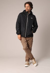 Man met krullend haar die een zwarte capuchonjack, beige cargobroek en tan sneakers draagt, met gekruiste benen staand tegen een plain achtergrond.