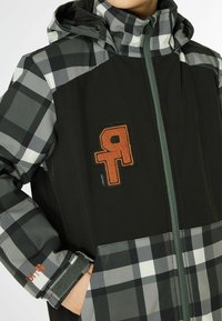 Veste à carreaux noir et gris avec capuche, arborant un logo orange brodé bien visible, des poignets élastiques et une fermeture éclair à l'avant.