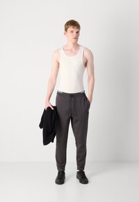Lys grå tanktop, mørkegrå joggebukser med snøre og sidelommer, sorte sneakers, sort jakke i hånden, ensfarvet baggrund.