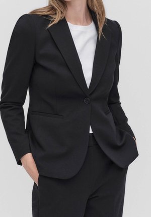 Kvinde iført en tætsiddende sort blazer over en hvid top med hænderne i lommerne mod en ensfarvet lys baggrund.