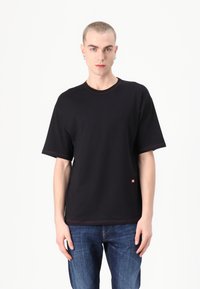 Czarny, oversize t-shirt z okrągłym dekoltem, krótkimi rękawami, kontrastującym czerwonym przeszyciem i małą metką z logo u dołu. Noszony z niebieskimi dżinsami.