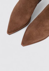 Bottines en daim brun avec des pointes effilées et une semelle en cuir lisse, présentant un design épuré et des coutures minimales.