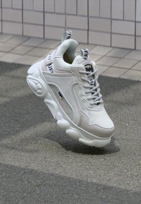 Weiße Sneaker mit einem Obermaterial aus Mesh und beigen Akzenten, runder Zehenpartie, dicke Sohle, auffälligen Schnürsenkeln und bedrucktem Branding auf der Zunge.