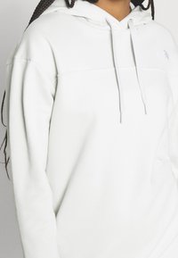 Sudadera blanca de tela suave, con capucha ajustable por cordón, puños acanalados y un pequeño logo en el pecho. Diseño simple y minimalista.