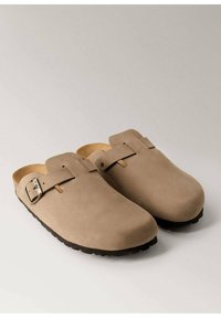Clogs din suede bej, cu o textură netedă, vârf rotund și un detaliu cu cataramă pe o parte. Talpă groasă din cauciuc pentru aderență.