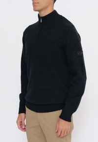 Uomo che indossa un maglione nero con mezza cerniera, polsini e orlo a coste, abbinato a pantaloni beige, in piedi davanti a uno sfondo bianco uniforme.