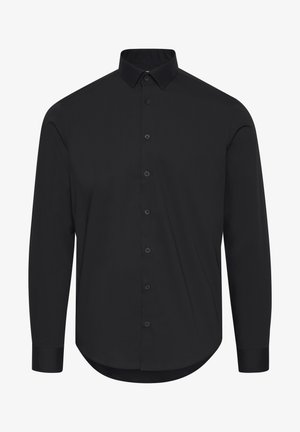 Mężczyzna w dopasowanej czarnej koszuli na guziki z długim rękawem i beżowych spodniach na jasnym, jednolitym tle.