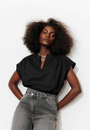 Femme confiante avec une coiffure afro naturelle, portant un chemisier noir à manches courtes et un jean gris taille haute, debout les mains sur les hanches.