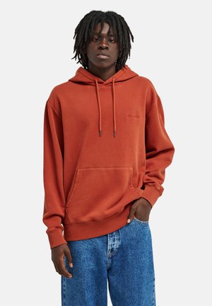 Junge Person mit dunkler Haut und Dreadlocks, die einen rostfarbenen Hoodie und lockere blaue Jeans trägt und mit einer Hand in der Tasche steht.