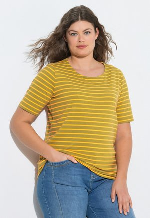 Femme aux longs cheveux ondulés portant un t-shirt à manches courtes rayé jaune et orange et un jean bleu, debout avec une main dans la poche.