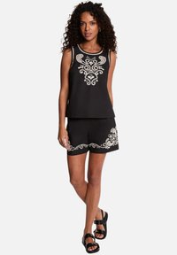 Mujer con top negro sin mangas y pantalones cortos con bordado decorativo blanco, combinados con sandalias negras abiertas, de pie sobre fondo blanco.