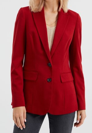 Vrouw draagt een nauwsluitend rood colbert met zwarte knopen over een beige top en donkere jeans, staand tegen een effen lichte achtergrond.