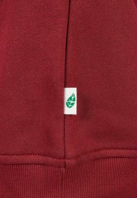 Burgundinės spalvos džemperio medžiaga su raštuotais rankogaliais. Išsiskiria balta etikete su žalia lapo logotipu, esančiu apatinėje siūlės pusėje. Lygus paviršius.