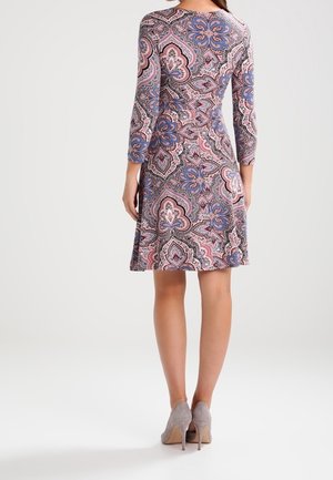 Femme debout portant une robe longueur genou avec des motifs complexes roses, bleus et noirs et des chaussures à talons gris, vue de dos.