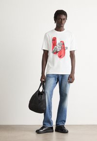 Mann in weißem T-Shirt mit roten Boxhandschuhen und Boxer-Print, blauen Jeans und schwarzen Sneakers, der eine schwarze Tasche vor einem weißen Hintergrund hält.