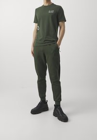 Zelený sportovní outfit sestávající z přiléhavého trička s krátkým rukávem a zúžených tepláků s černými postranními pruhy. Celý vzhled doplňují černé tenisky.