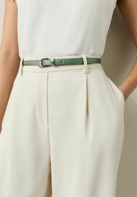Lys grøn læderbælte med sølvhardware båret over cremefarvede skræddersyede bukser, der har plisseringer og sidelommer.