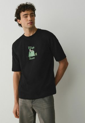 Młody mężczyzna z kręconymi włosami, ubrany w czarny t-shirt z zielonym nadrukiem Beagle Scouts oraz szare jeansy, stojący na jednolitym tle.