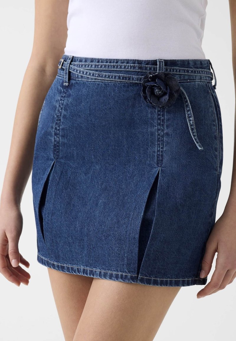 Femme portant une mini-jupe en jean bleu avec des plis devant et un accessoire de ceinture en denim en forme de fleur, associée à un haut blanc côtelé.