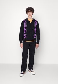 Cárdigan negro con patrón morado, camiseta naranja debajo, pantalones negros y zapatillas multicolores. Tela suave, delantero con botones, puños acanalados.