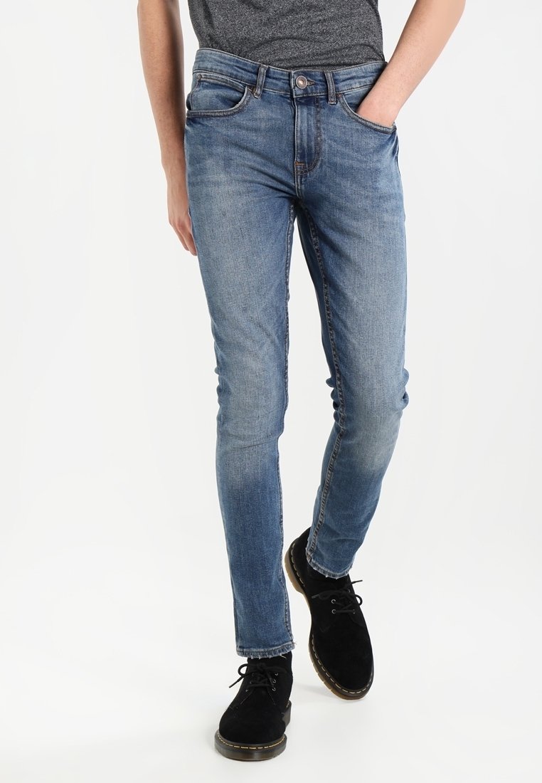 Vaqueros de mezclilla azul con un diseño ajustado, que presentan un estilo clásico de cinco bolsillos y un ligero desvanecimiento; combinados con zapatos negros.