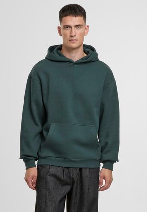 Mørkegrøn sweatshirt med kængurulomme, justerbar snørelys og ribbede manchetter, lavet af blødt materiale. Model iført den.