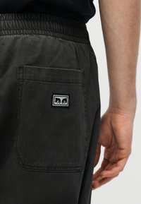 Fekete nadrág közeli képe, rugalmas derékpánttal, hátul egy zsebbel, amelyen egy kis téglalap alakú folt látható két stilizált fekete állatarccal.