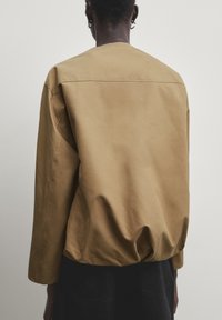Veste marron en mélange de coton avec une coupe ample, présentant un col rond et un ourlet froncé. Texture lisse avec des détails de coutures minimaux.