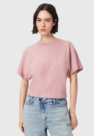 Ung kvinde med kort mørkt hår iført en lyserød kortærmet top og højtaljede lyseblå jeans mod en ensfarvet baggrund.
