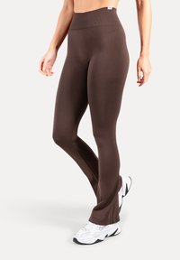 Braune Leggings mit hoher Taille, glatter Textur und ausgestelltem Beinabschluss. Das Modell trägt weiße Sportschuhe als Kontrast.