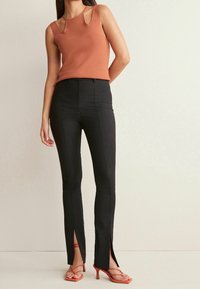 Rust-kleurig mouwloos top met sleutelgat uitsparingen, gecombineerd met hoge taille zwarte broek met zijsplitten en rode bandjeshakken.