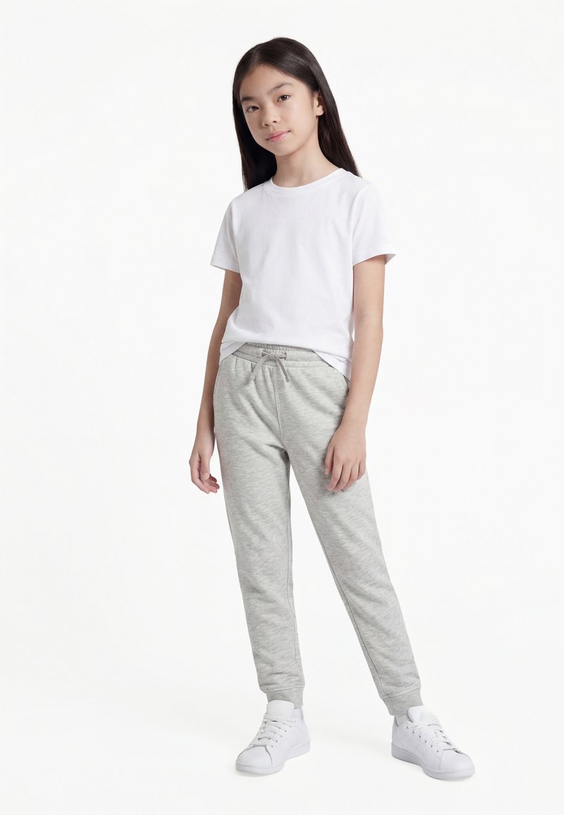 Djevojka stoji, nosi bijelu majicu kratkih rukava, svijetlosive trenirke s vrpcom za vezanje i bijele tenisice, na jednobojnoj pozadini.