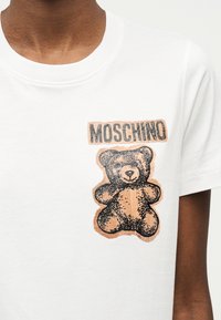 Camiseta blanca con un gráfico de un oso de peluche marrón y la palabra "MOSCHINO" impresa arriba, en el lado izquierdo del pecho.
