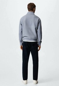 Felpa grigia con colletto alto, polsini e vita a costine, abbinata a pantaloni scuri e sneakers bianche, vista da dietro.