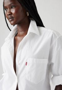 Camicia bianca con bottoni, caratterizzata da una tasca sul petto e un'etichetta rossa del marchio. Il tessuto appare liscio, con un colletto classico e maniche arrotolate.