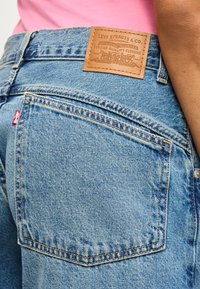 Κοντινό πλάνο της πίσω τσέπης μπλε τζιν Levi's με δερμάτινο ετικέτα μάρκας και κόκκινη ετικέτα λογότυπου, φοράει ροζ πουκάμισο.