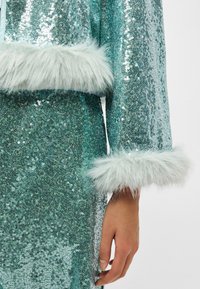 Roupa em turquesa com lantejoulas, com punhos de pelúcia falsa em azul claro suave. O tecido tem uma textura brilhante, captando a luz de forma eficaz.