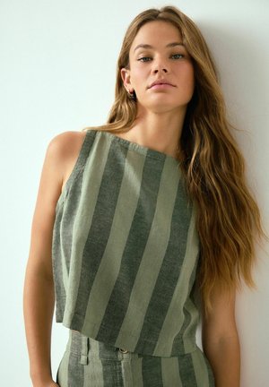 Frau mit langen, welligen Haaren, die ein ärmelloses Oberteil mit grünen und grauen vertikalen Streifen sowie passende hochtaillierte Hosen trägt und vor einer weißen Wand steht.