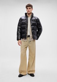 Μαύρο γυαλιστερό τζάκετ με επένδυση και ψηλό γιακά, σε συνδυασμό με ένα ανοιχτό ριγέ πουκάμισο και μπεζ παντελόνι με φαρδιά γραμμή. Μαύρα παπούτσια ολοκληρώνουν το ντύσιμο.