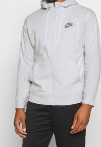 Svetle šedá mikina na zip s kapucí na šňůrku, předními kapsami a černým logem Nike. Měkká tkanina s žebrovanými manžetami a lemem.