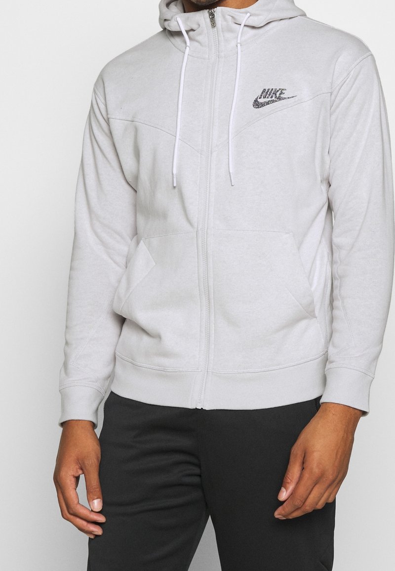 Svetle šedá mikina na zip s kapucí na šňůrku, předními kapsami a černým logem Nike. Měkká tkanina s žebrovanými manžetami a lemem.