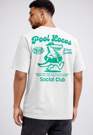 Man wearing white T-shirt with turquoise graphic of cartoon crocodile in inner tube and text promoting a Sunday pool social club.
