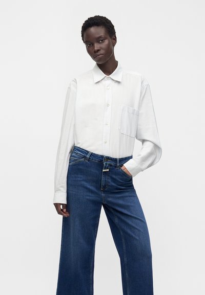 Femme portant une chemise blanche à boutons avec une poche poitrine et un jean large bleu foncé taille haute, debout sur un fond uni.