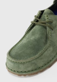 Grüne Wildleder-Mokassinschuhe mit Steppnähten, Schnürsenkeln und einer durchsichtigen Gummisohle auf weißem Hintergrund.