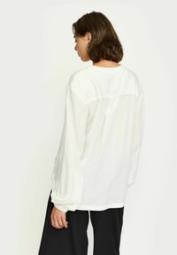 Blusa bianca a maniche lunghe con vestibilità rilassata, dotata di un yoke sul retro e una sottile texture. Abbinata a pantaloni neri larghi.