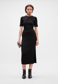 Rochie tricotată neagră, cu mâneci scurte, fustă până la genunchi cu o fente laterală. Modelul poartă bocanci negri până la gleznă și ține o geantă mică neagră.