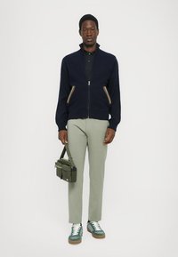 Marinblå zip-up tröja med flerfärgade ränder på fickorna, ljusgrå byxor och gröna sneakers. Modellen håller en olivgrön budväska.