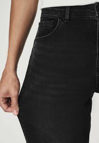 Gros plan d'une personne portant un jean noir, tirant légèrement le tissu loin de la cuisse pour montrer l'élasticité.