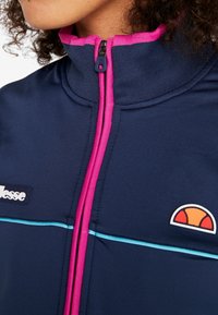 Marinblå zip-up jacka med hög krage, rosa dragkedja och ljusblå rand. Logotyp på vänster sida. Texturerat, atletiskt tyg.